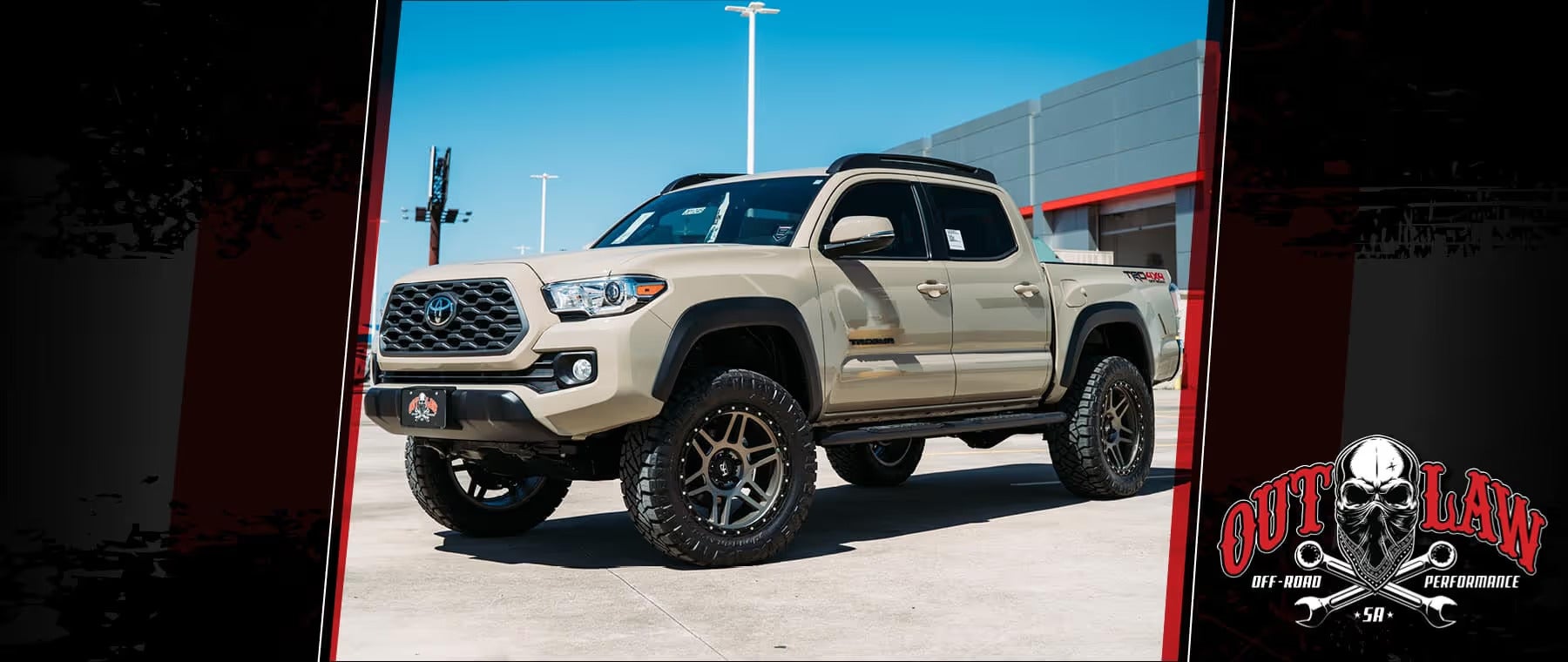 Lifted beige Toyota Tacoma truck with large off-road tires parked outside a building under a clear blue sky.